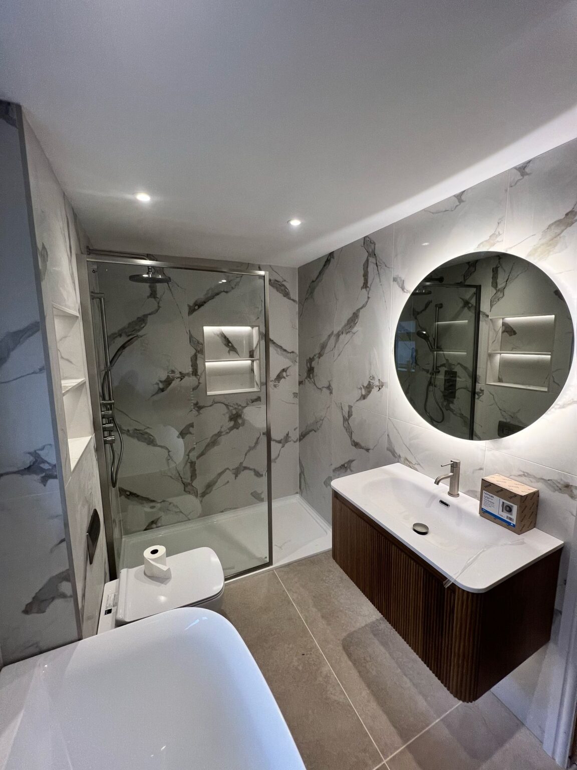 Modern bathroom with marble-effect tiles, walk-in shower, round illuminated mirror, and floating vanity unit