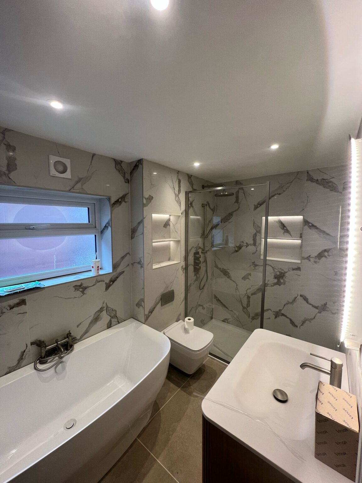 Modern bathroom with marble-effect tiles, walk-in glass shower, freestanding bathtub, and illuminated vanity mirror