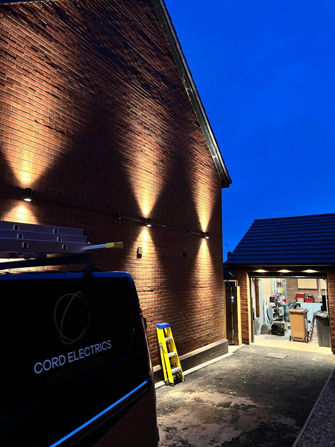 Cord Electrics van parked beside a brick property with newly installed exterior wall lights illuminating the pathway at dusk